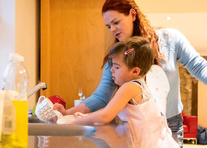 child in dress washes dishes with red-headed adult